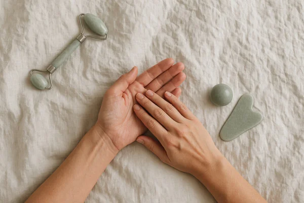 Relaxed hands resting on soft linen beside a jade roller, a massage ball and a gua sha stone